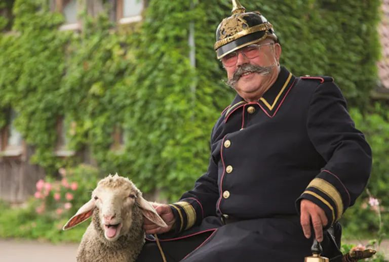 Der Büttel in dunkelblauer Uniform mit Pickelhaube sitzt lachend neben einem Schaf