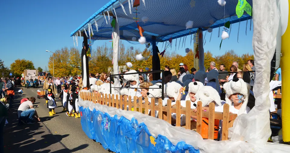 Geschmückter Festwagen im Umzug: Kinder als Eisbären unter einem blauen Netzdach, daneben Kinder als Pinguine