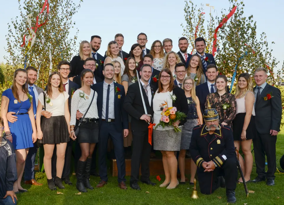 Gruppenbild auf einer Wiese vor Bäumchen mit bunten Bändern: junge Frauen und Männer in festlicher Kleidung mit Anstecksträußchen, in der Mitte ein Mann mit einem Degen, an dem ein rotes Band hängt, und eine Frau mit einem bunten Blumenstrauß; vorn kniet der Büttel in dunkelblauer Uniform mit Pickelhaube, neben ihm steht eine Glocke im Gras.