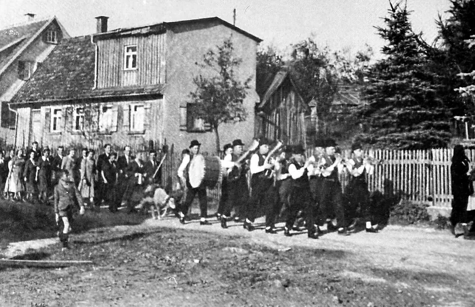 Schwarz-Weiß-Aufnahme: Eine Blaskapelle in Tracht und mit dunklen Hüten marschiert über eine Dorfstraße, bei ihr ein Schaf; am Straßenrand stehen Zuschauerinnen und Zuschauer vor alten Häusern.